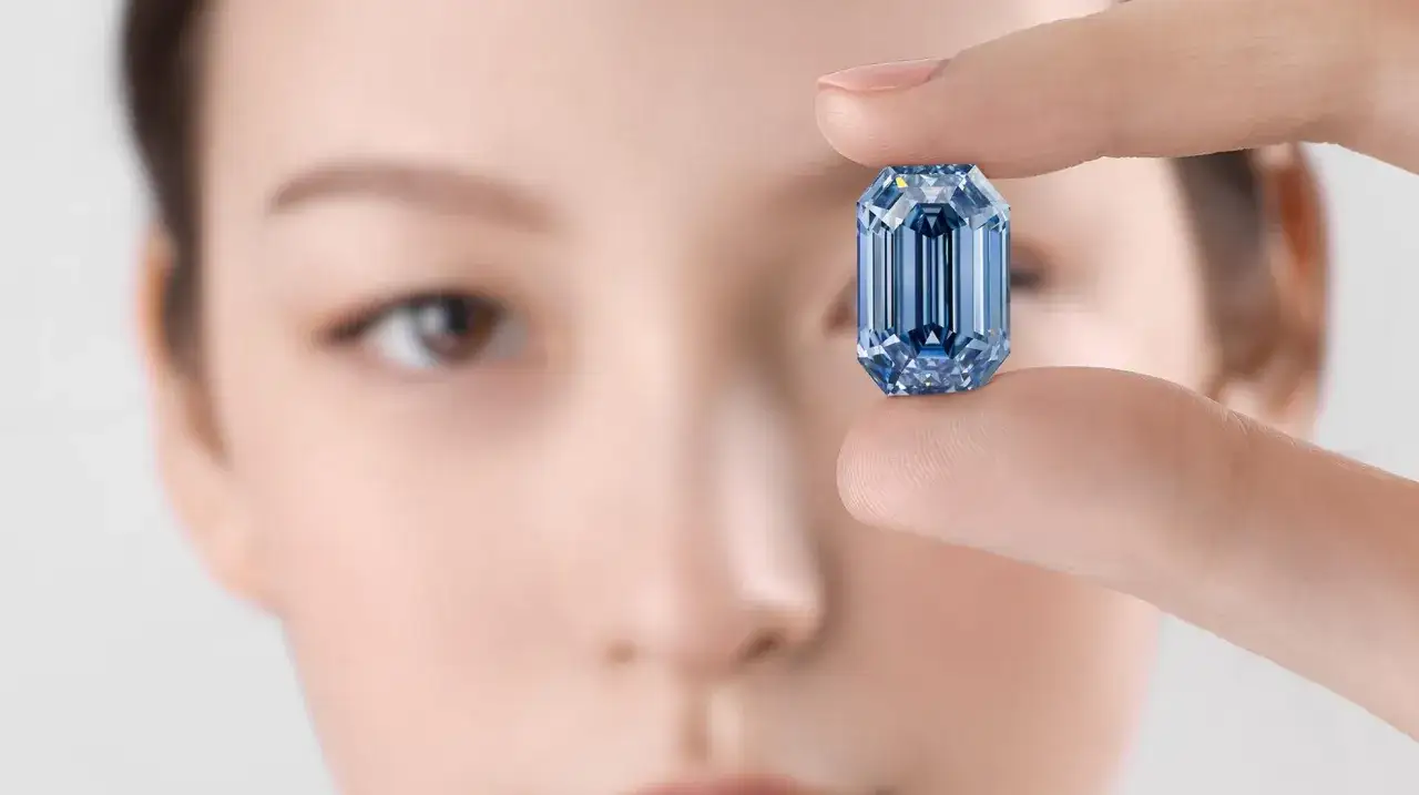 Dłoń trzyma błyszczący, niebieski diament o szlifie emerald. W tle rozmyta twarz kobiety.