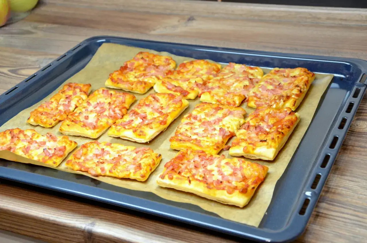 Chrupiące ciasto francuskie z serem i kawałkami szynki, ułożone na blasze wyłożonej papierem do pieczenia.