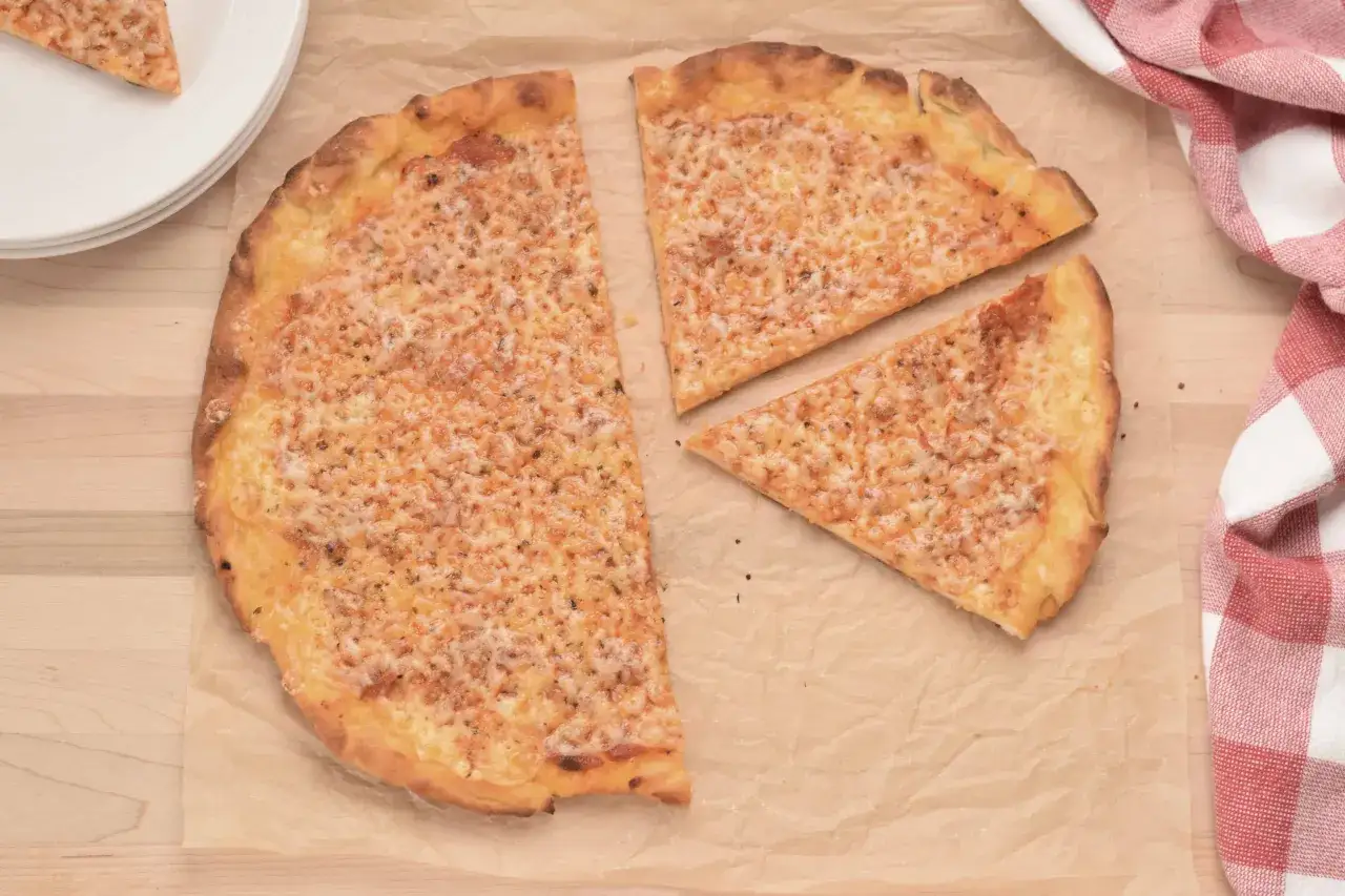 Pyszna pizza bez drożdży, posypana obficie serem, leży na papierze do pieczenia. Obok leżą talerze i czerwono-biała ściereczka.