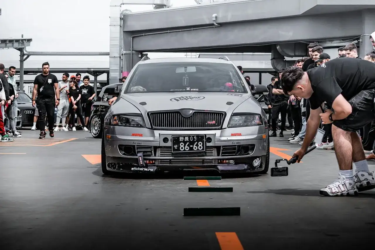 Mężczyzna z kamerą ustawia sprzęt do nagrywania **tuning**u srebrnego samochodu na zlocie fan&oacute;w motoryzacji.