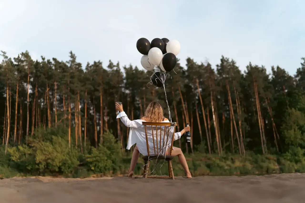 Kobieta świętuje sesję urodzinową w plenerze, trzymając kieliszek i butelkę szampana, otoczona czarno-białymi balonami.