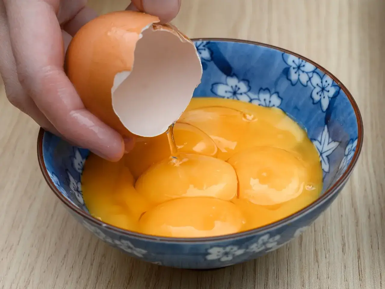 Ręka wylewa ż&oacute;łtka z poł&oacute;wki skorupki do niebieskiej miseczki. Wiele ż&oacute;łtek czeka na pomysły, co można zrobić z ż&oacute;łtek.