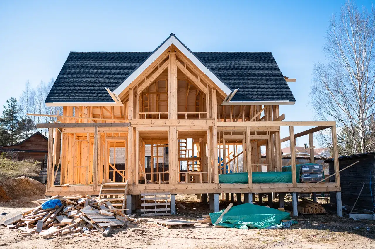 Budowa domu kanadyjskiego w stanie surowym. Drewniana konstrukcja, dach pokryty gontem, materiały budowlane na placu.