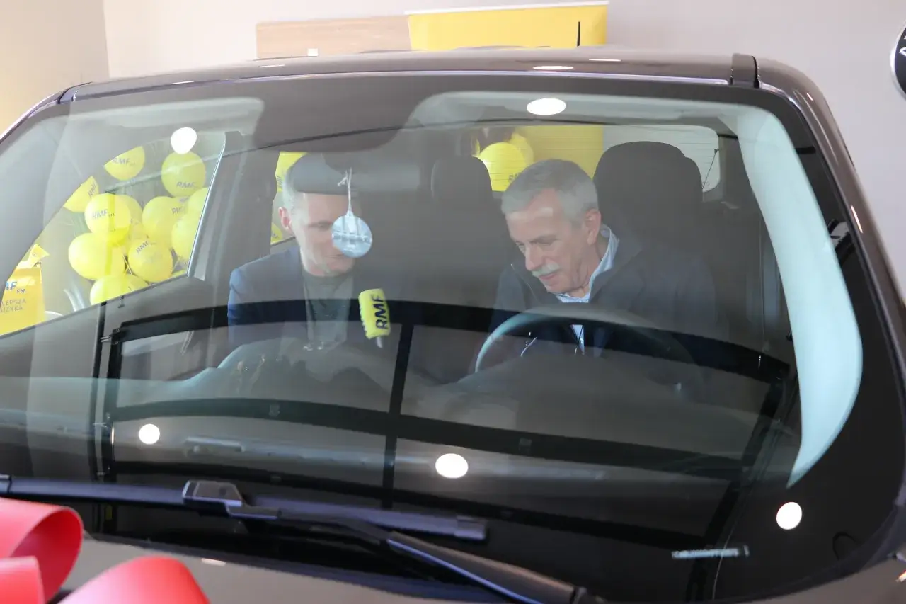 Dwóch mężczyzn siedzi w samochodzie, za kierownicą i obok. W tle widać żółte balony z logo RMF FM. To może być finał loterii RMF.