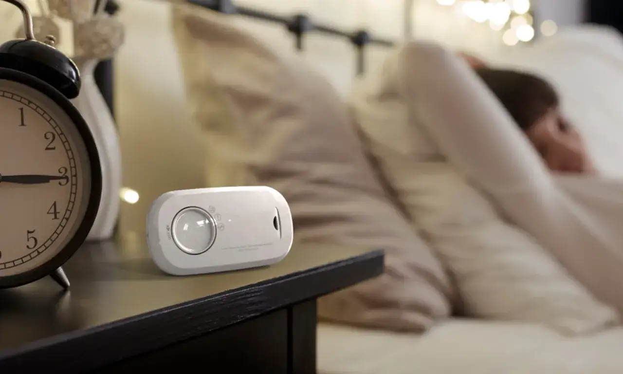 Na stoliku nocnym obok budzika stoi czujnik gazu. Czy czujnik gazu reaguje na parę wodną? W tle śpi osoba.