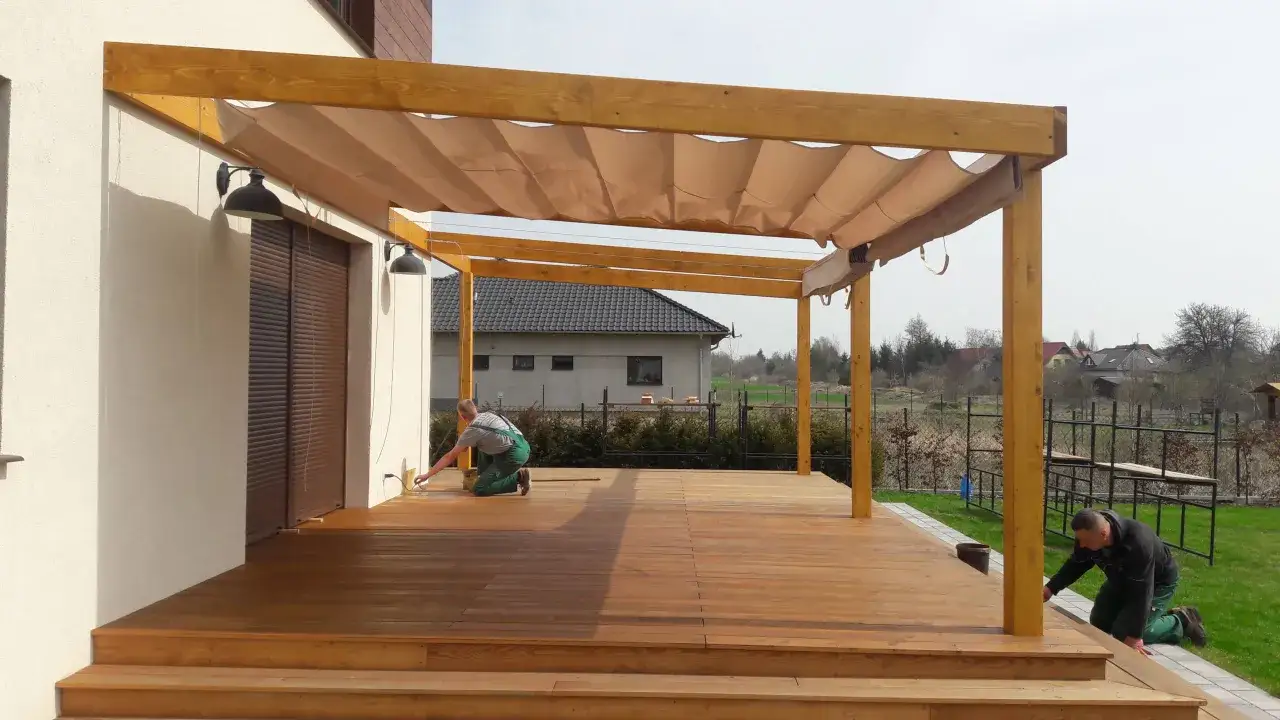 Drewniane tarasy na słupach z zadaszeniem, kt&oacute;re właśnie powstają. Dw&oacute;ch mężczyzn pracuje nad ich wykończeniem.