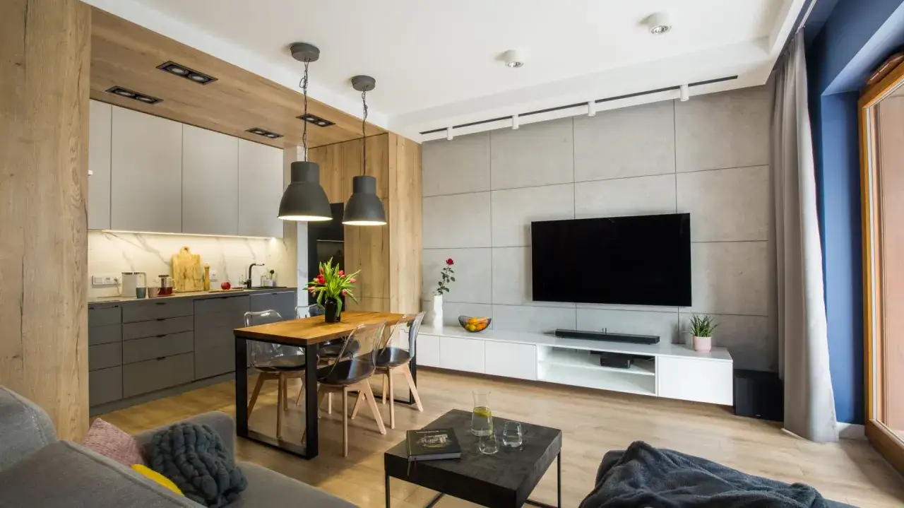 Nowoczesny salon z aneksem kuchennym. Betonowe płytki na ścianie z telewizorem, drewniane elementy i szare meble kuchenne. Beton w salonie inspiracje.