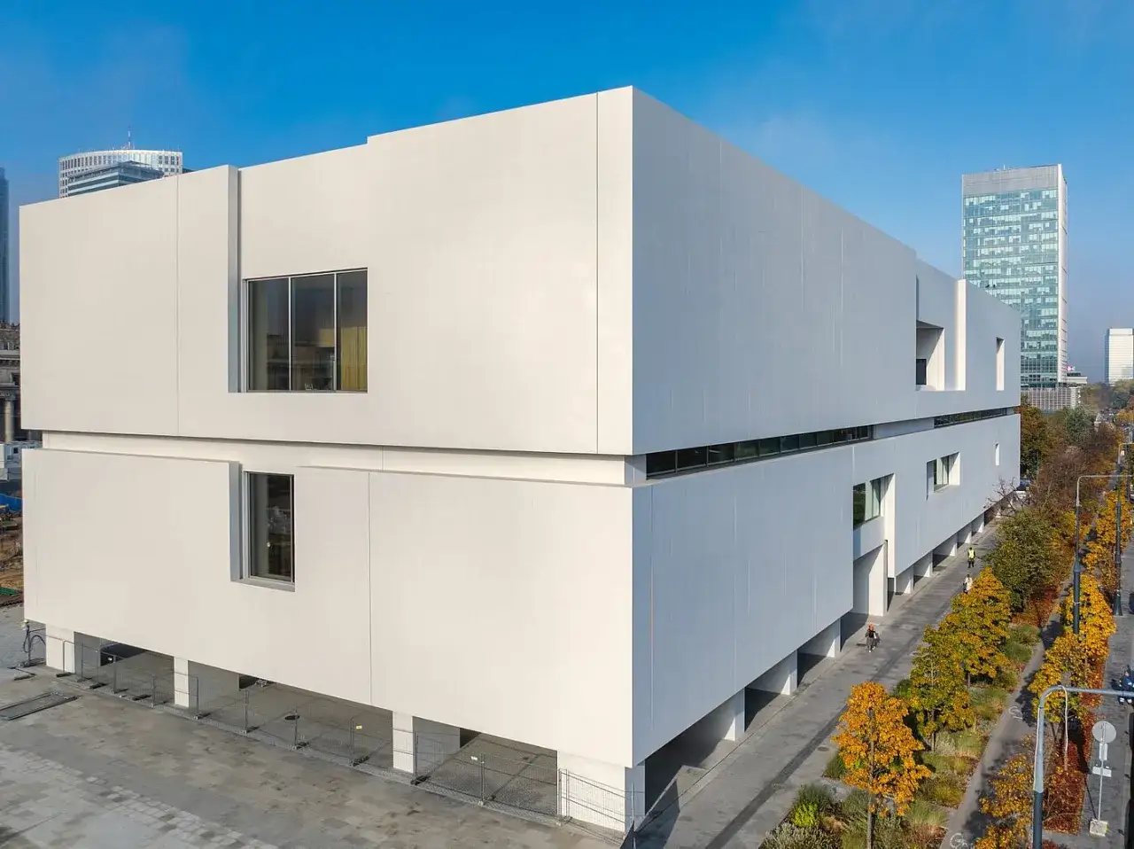 Nowe muzeum sztuki nowoczesnej w Warszawie, biały, geometryczny budynek z dużymi oknami, otoczony jesienną zielenią i nowoczesną architekturą miejską.