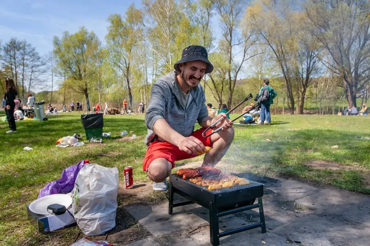 Mężczyzna w czerwonych spodenkach i kapeluszu grilluje kiełbaski i ziemniaki na trawie. W tle widać ludzi odpoczywających na pikniku, idealne miejsce gdzie na grilla.