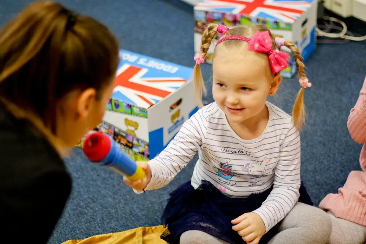 Uśmiechnięta dziewczynka z kucykami podaje mikrofon, pokonując barierę językową u dziecka.