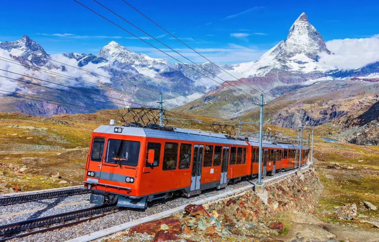 Pomarańczowy pociąg jedzie przez malownicze góry w Szwajcarii, z ośnieżonym szczytem Matterhorn w tle.