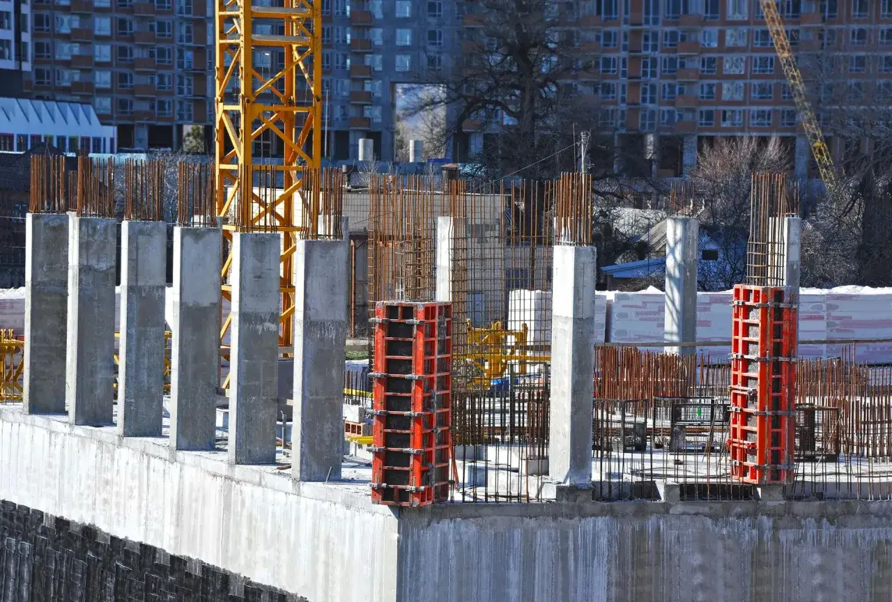 Budowa postępuje, widać betonowe słupy i zbrojenie, które jest kluczowe, by wiedzieć jak krecic zbrojenie. W tle budynek i dźwig.