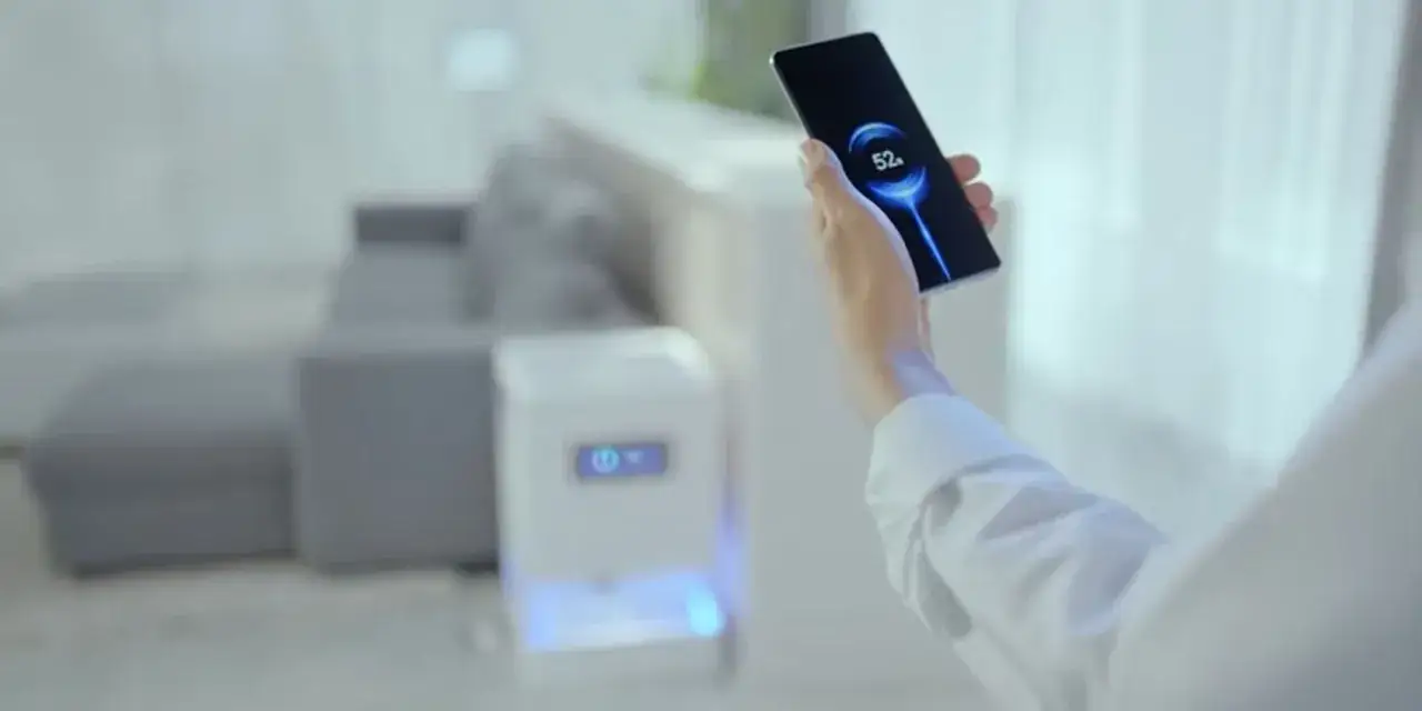 Dłoń trzyma smartfon Xiaomi z ikoną ładowania 52%. W tle widać meble i białe urządzenie z niebieskim podświetleniem.