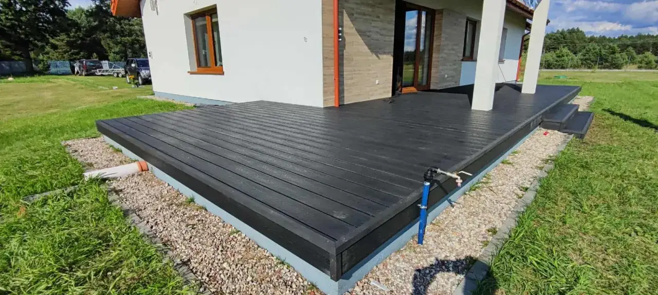 Nowy taras z czarnego kompozytu otacza dom w kształcie litery L, tworząc przestrzeń do wypoczynku.