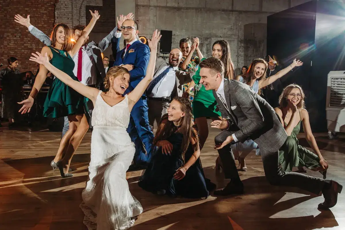 Radosne tańce na weselu! Goście w r&oacute;żnorodnych strojach bawią się na parkiecie, unosząc ręce w geście radości.