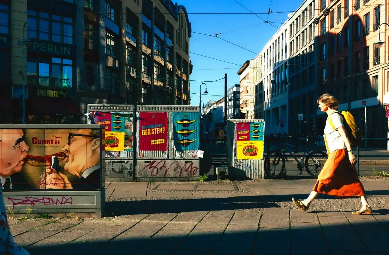 Kobieta z plecakiem idzie ulicą w Berlinie. W tle kolorowe plakaty, reklamy i budynki. To klasyczna fotografia uliczna.