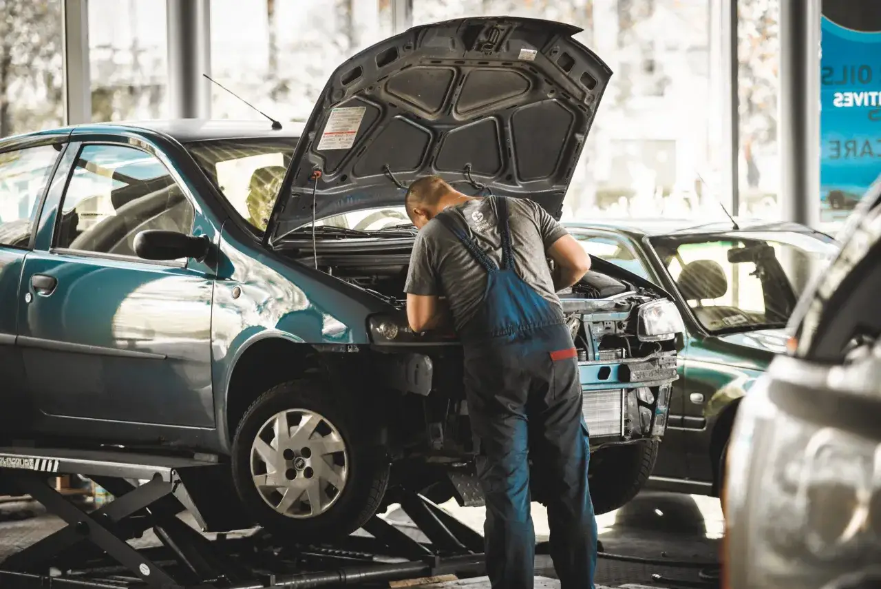 Mężczyzna w warsztacie samochodowym, uczący się mechaniki. Podniesiony samochód, otwarty przód, praca przy silniku. Kurs mechanika samochodowego dla dorosłych.