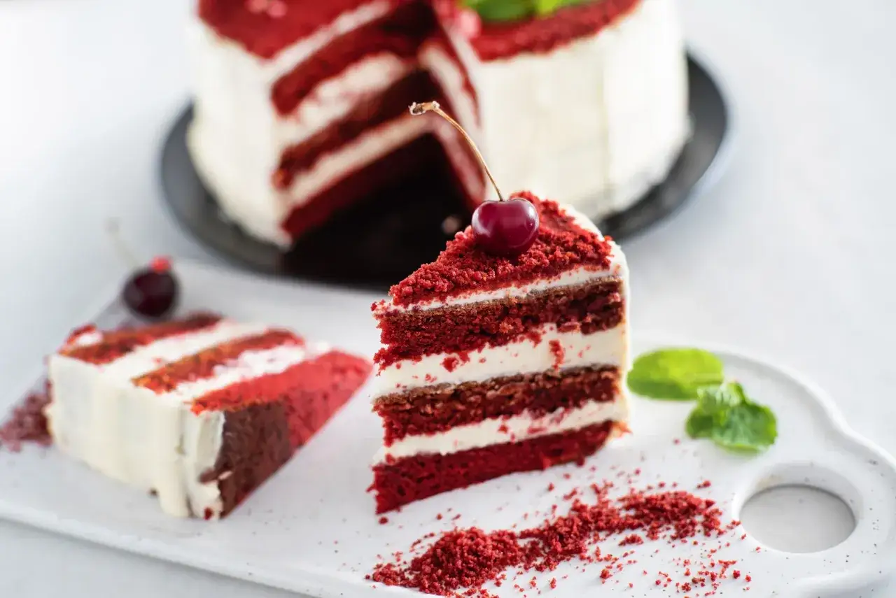 Kawałek tortu red velvet z kremem i wiśnią na wierzchu, obok leży drugi kawałek i listki mięty.