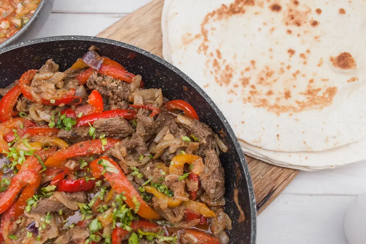 Gorące fajitas co to? Aromatyczne mięso z papryką i cebulą na patelni, obok stos tortilli.