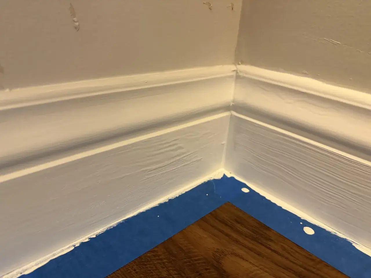 Malowanie listew przypodłogowych: biała farba na drewnie, zabezpieczone niebieską taśmą.