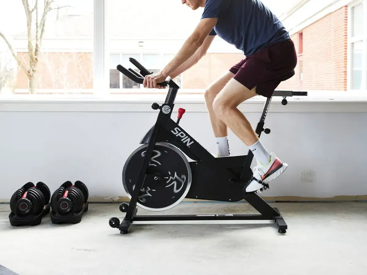 Intensywny trening cardio na rowerze stacjonarnym. Obok stoją regulowane hantle, gotowe do dalszych ćwiczeń.