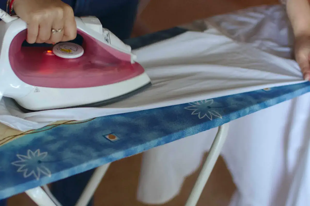 Dłoń z żelazkiem prasuje biały rękaw koszuli na desce.