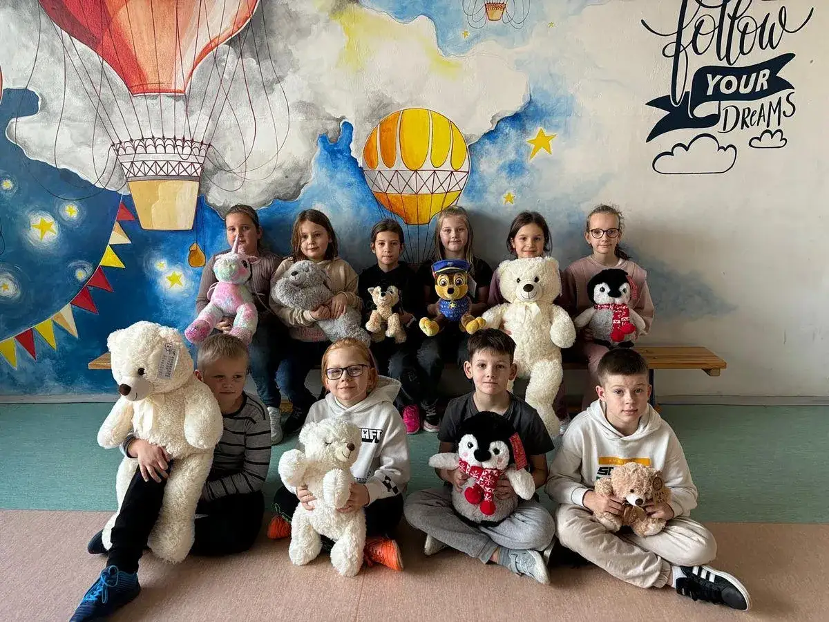 Dzieci świętują dzień misia, pozując z pluszakami na tle malowidła z balonami i gwiazdami.
