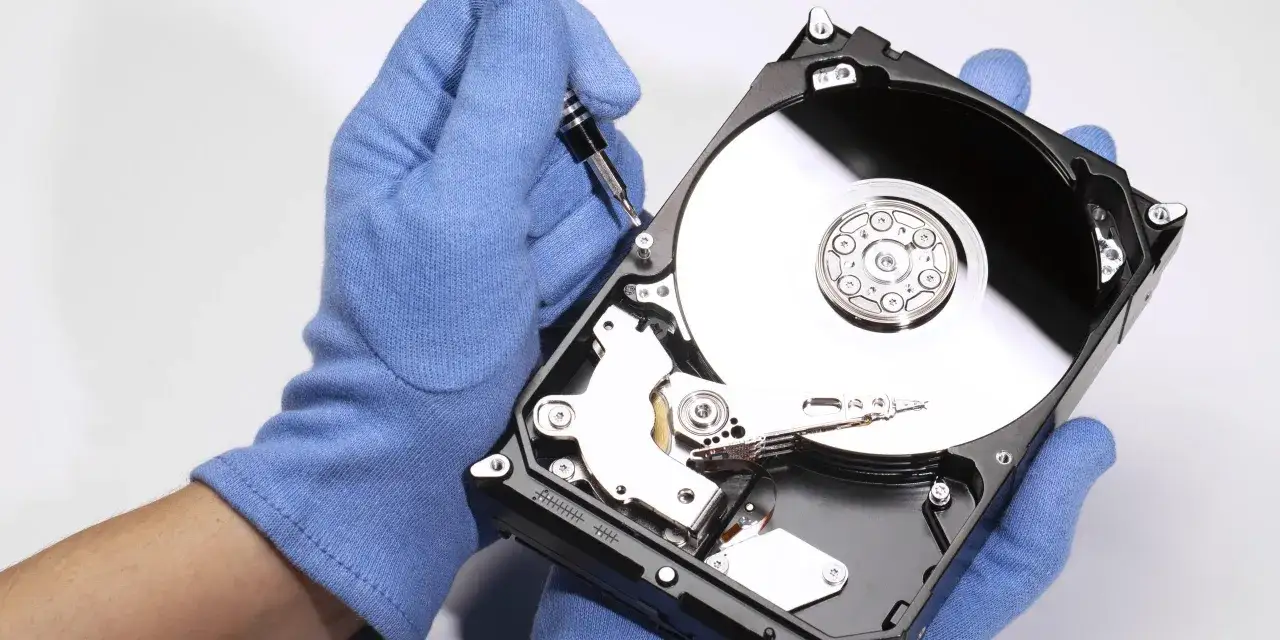 Ręce w niebieskich rękawiczkach składają dysk twardy. Widoczny błyszczący talerz i mechanizm głowicy, jakby zbierano crystal disk info.