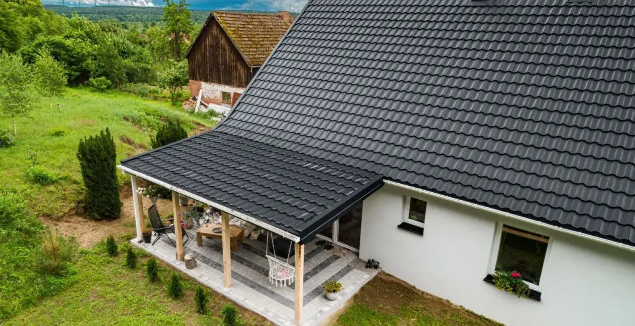 Nowy dach z czarnej blachy nad tarasem, idealny do wymiany starej pokrywy. Wokół zielony ogród i wiejski krajobraz.