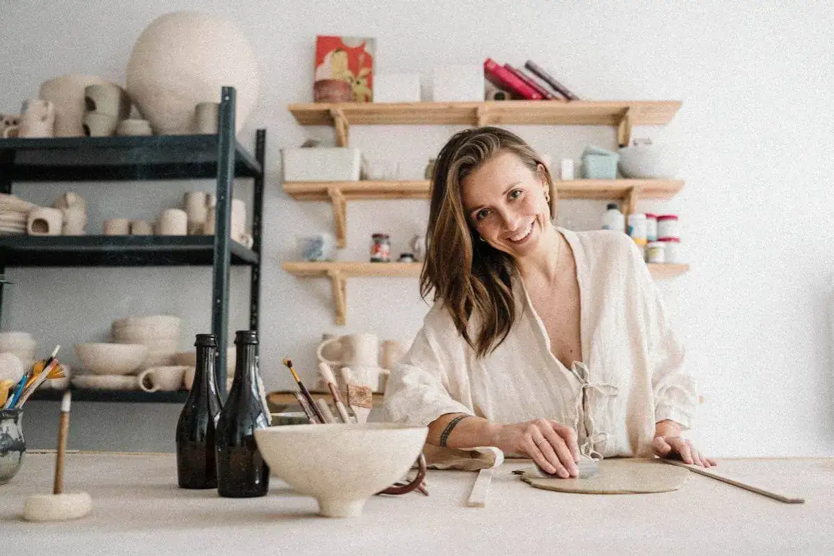 Uśmiechnięta kobieta podczas zajęć z ceramiki, tworzy dzieło z gliny. W tle p&oacute;łki z naczyniami i narzędziami.