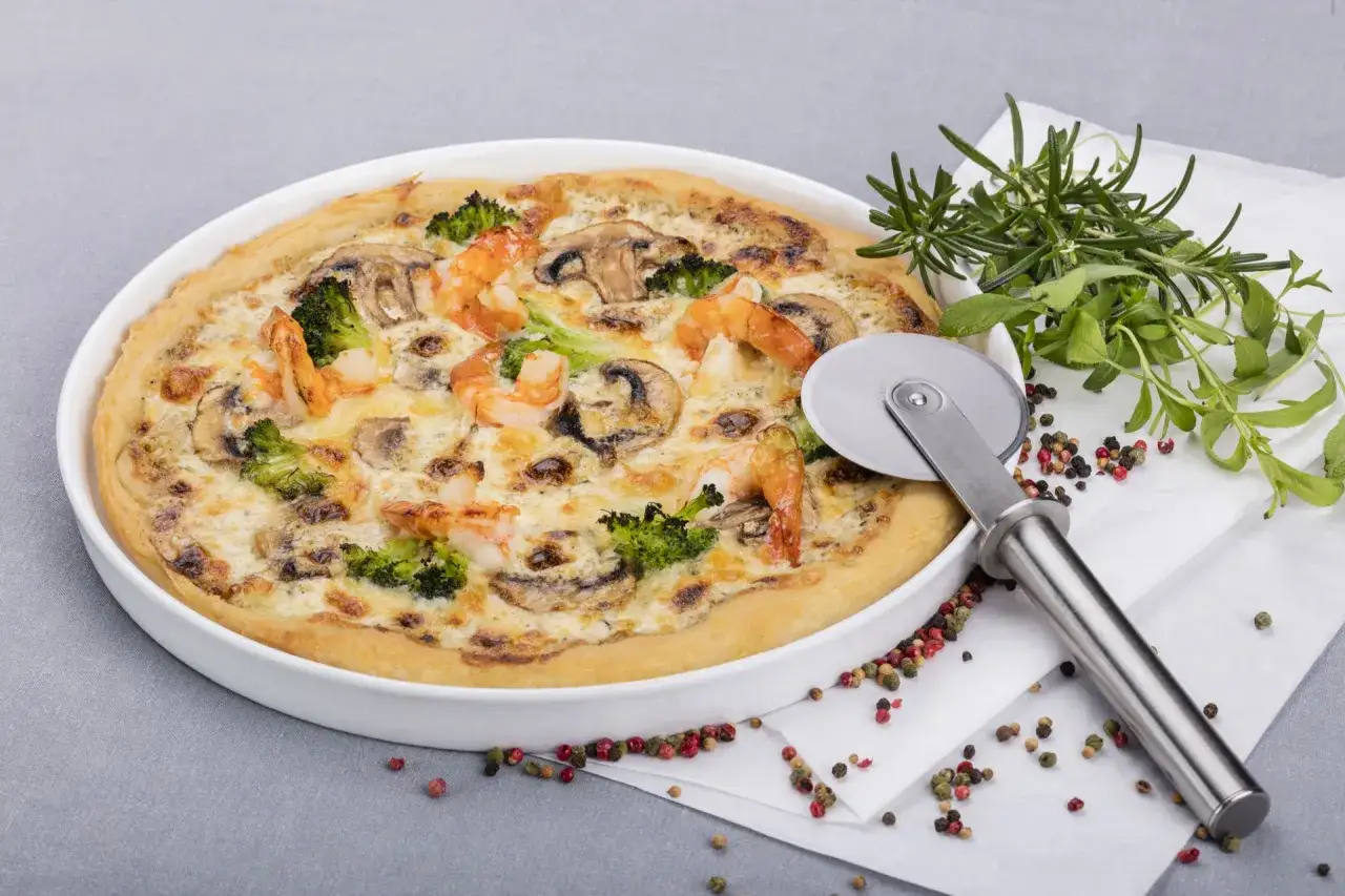 Pyszna pizza z krewetkami, brokułami i pieczarkami w białym naczyniu, obok nóż do pizzy i świeże zioła.