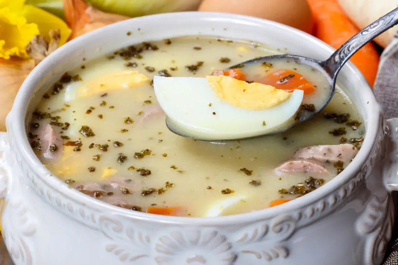 Miseczka żurku z jajkiem, kiełbasą i marchewką, na łyżce kawałek jajka.