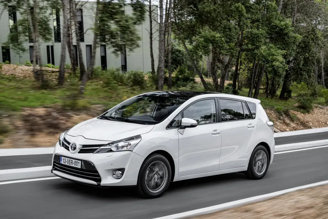 Biała Toyota Verso z czarnym dachem jedzie po drodze wśród drzew.