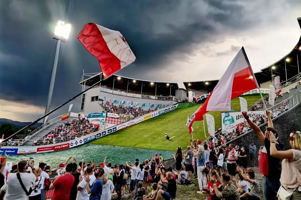 Tłumy kibiców z polskimi flagami na trybunach i na trawie, dopingujące skoczka podczas zawodów Pucharu Świata w Wiśle.