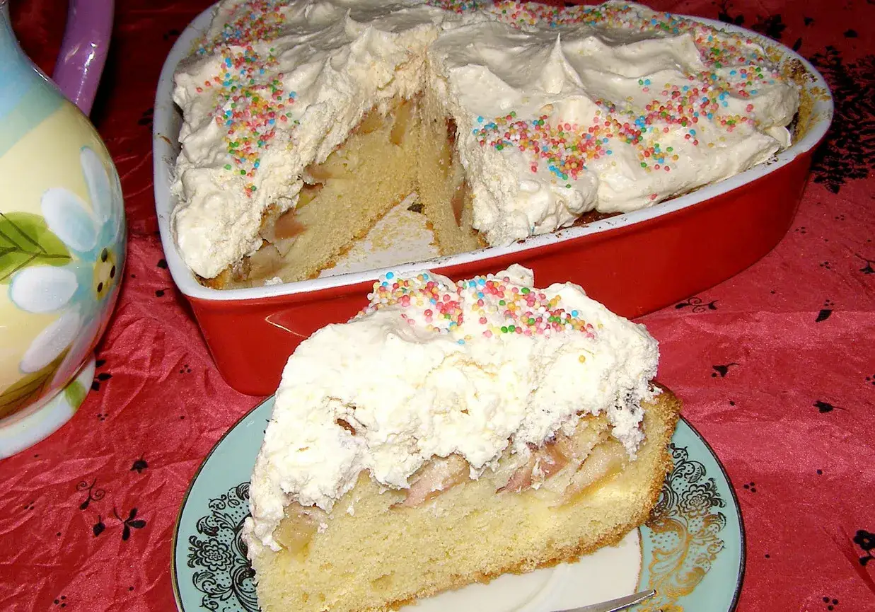 Pyszne ciasto z mąki krupczatki, przełożone jabłkami i udekorowane bitą śmietaną oraz kolorową posypką. Idealne na deser!