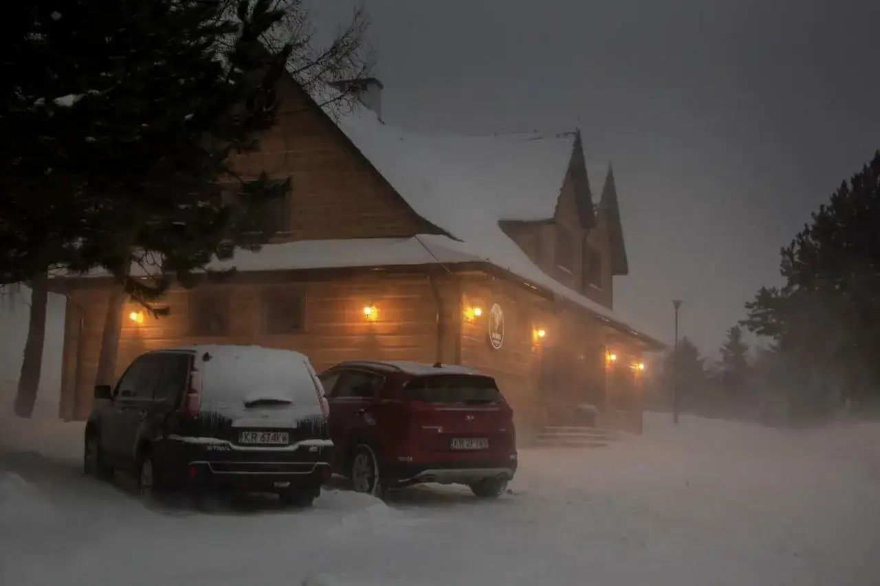 Zadymiona, zimowa noc. Drewniana bac&oacute;wka na zadzielu, oświetlona ciepłym światłem, tonie w śnieżnej zamieci. Dwa samochody czekają na gości.