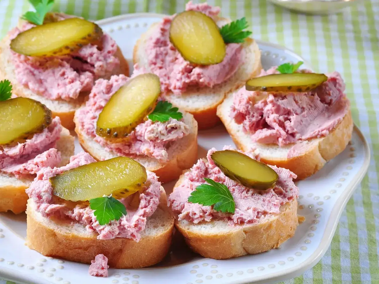 Kromki chleba z pastą z szynki, ozdobione plasterkami ogórka kiszonego i natką pietruszki, wyglądają apetycznie.