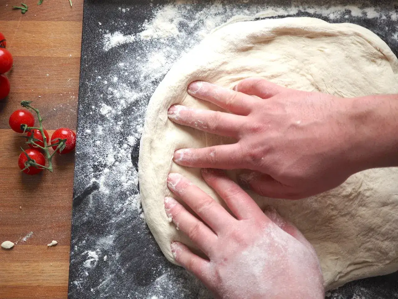 Ręce formują ciasto na pizzę, przygotowując je do pieczenia. To pierwszy krok, jak zrobić pizzę, obok świeżych pomidork&oacute;w.