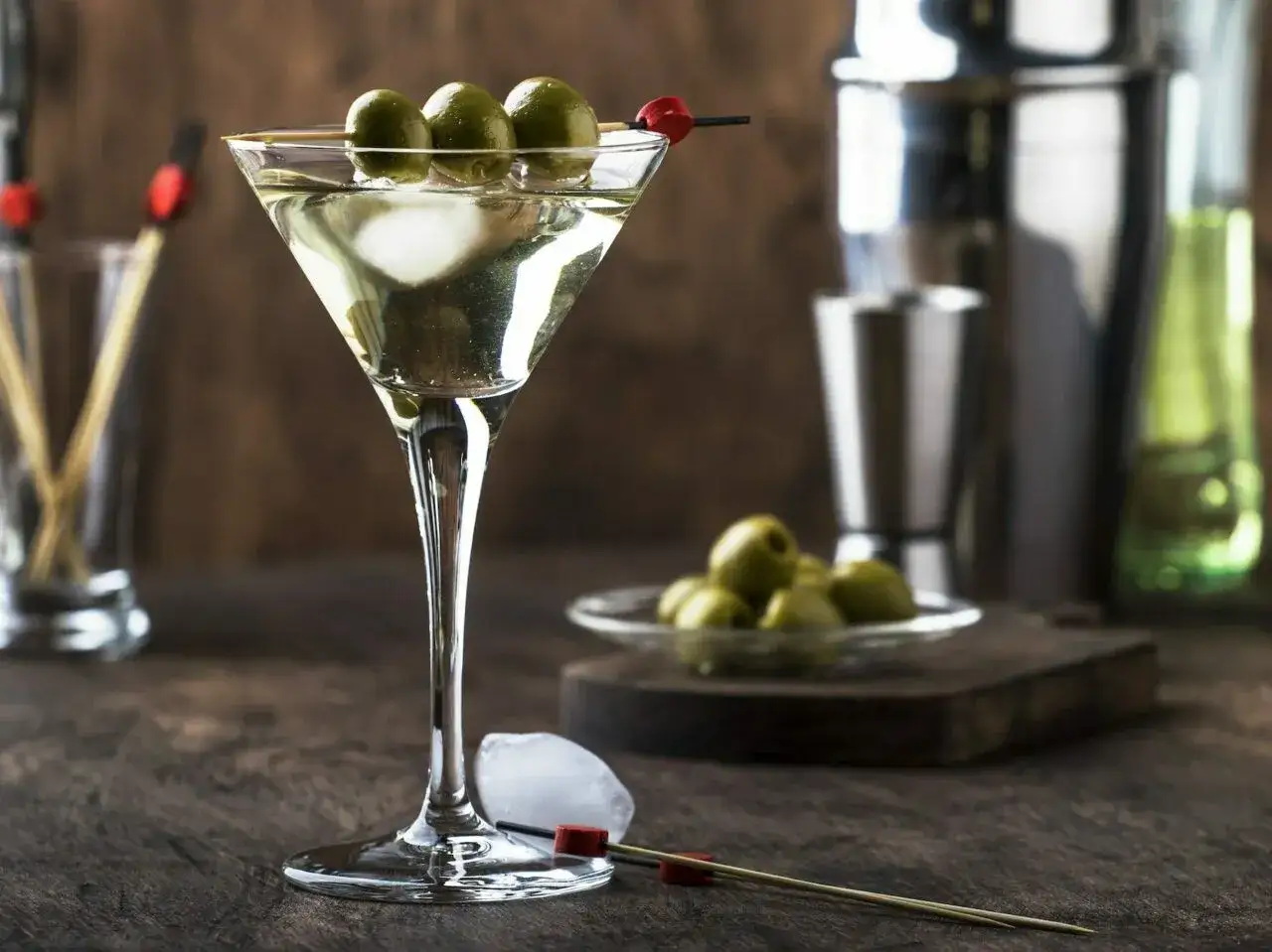 Elegancki kieliszek z drinkiem Martini Bianco, ozdobiony trzema oliwkami na patyczku. Obok kostki lodu i kolejny patyczek.