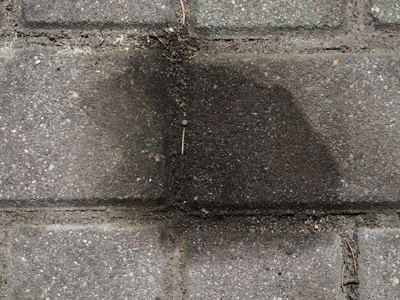 Stare plamy z oleju na kostce brukowej tworzą ciemny, rozlany kształt. Widać drobne kamyczki i piasek wtopione w nawierzchnię.