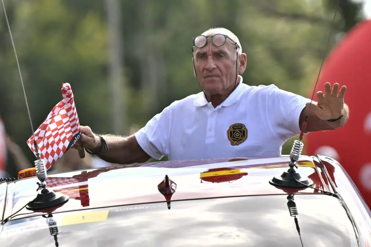 Starszy mężczyzna w okularach, machający chorągiewką w kratkę, jakby chciał powiedzieć: 