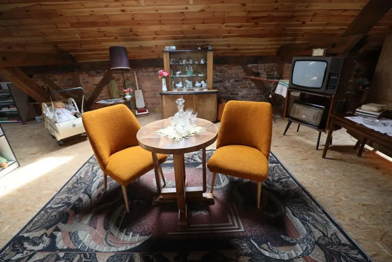 Pokój lata 90. z dwoma pomarańczowymi fotelami, stolikiem z karafką i kieliszkami, starym telewizorem i wózkiem dla lalki.