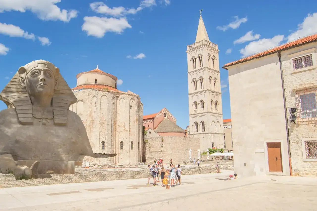 Zadar co zobaczyć: Sfinks, romańska bazylika i dzwonnica Katedry św. Anastazji. Turyści spacerują po placu.