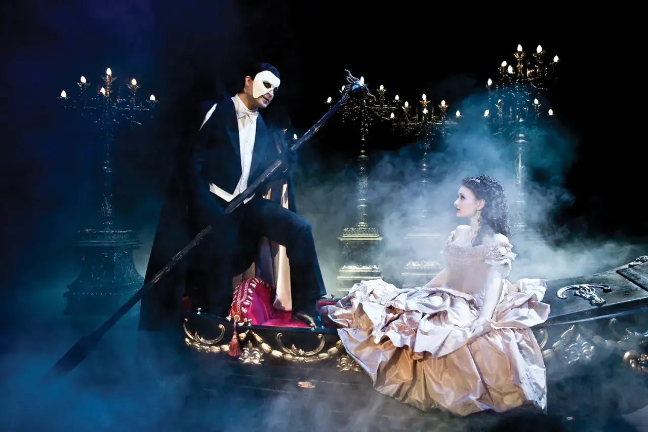 Upiór w operze w Warszawie: tajemnicza postać w masce i piękna Christine na łodzi, otoczeni mgłą i blaskiem świec.