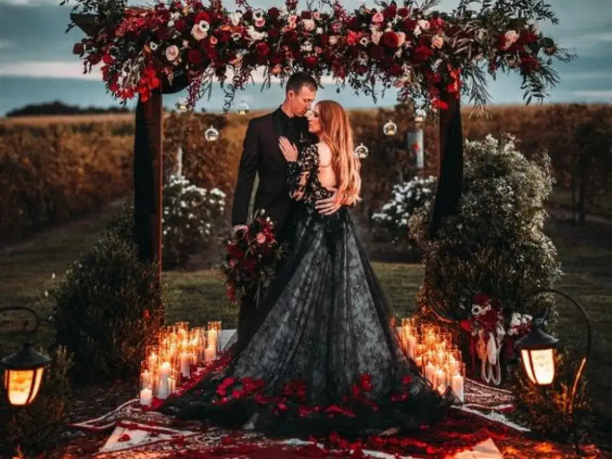 Para młoda pod łukiem z kwiatów. Ona w zjawiskowej czarnej sukni ślubnej z koronką i czerwonymi akcentami. Wokół mnóstwo świec i płatków róż.