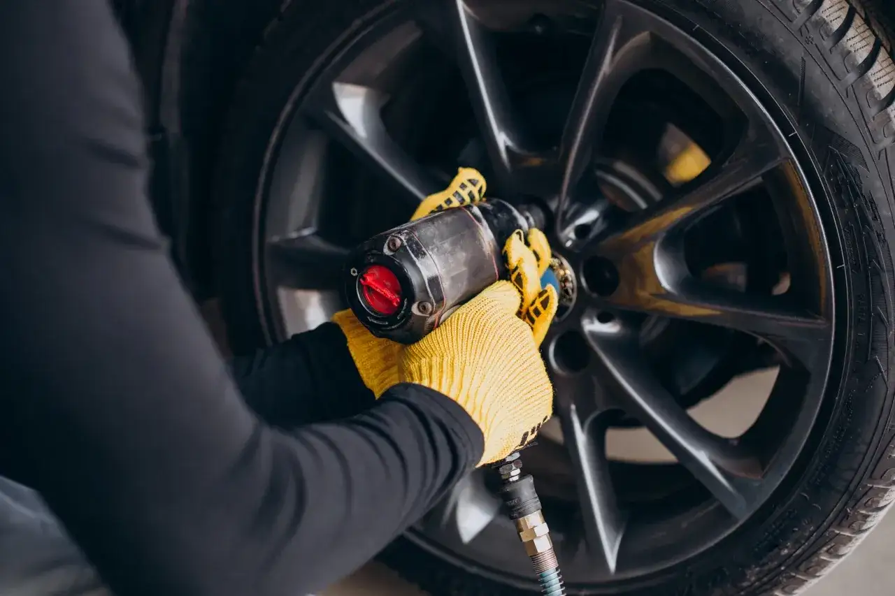 Mechanik w żółtych rękawicach używa klucza pneumatycznego do odkręcania śrub w feldze. Dowiedz się, ile kosztuje prostowanie felg.
