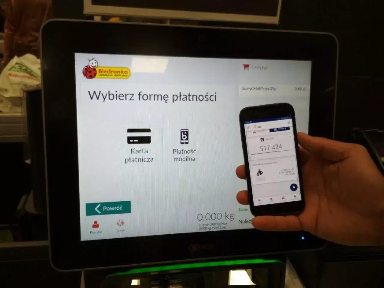 Na ekranie kasy Biedronki wybór formy płatności. Obok ekranu trzymany jest smartfon z widocznym kodem BLIK. Czy w Rossmannie można płacić Blikiem? Tutaj tak!