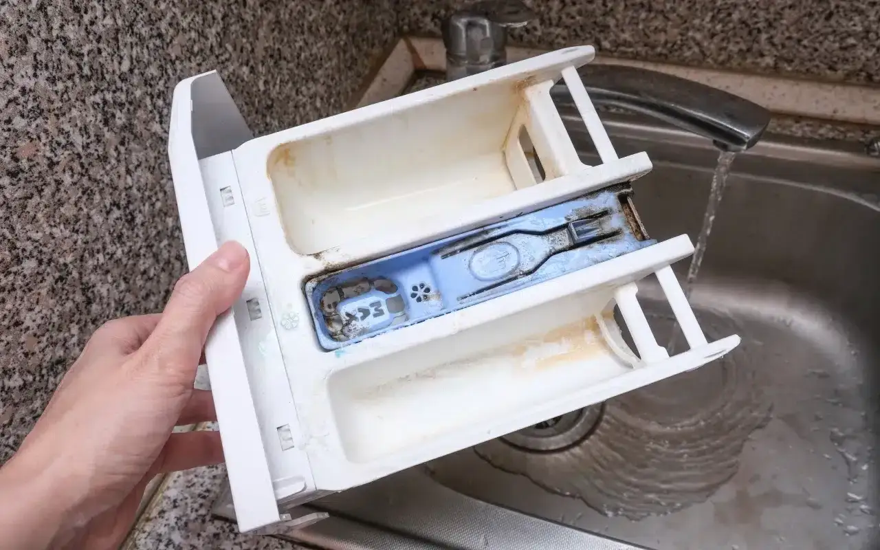 Brudny dozownik pralki, kt&oacute;ry nie pobiera płynu do płukania. Widać osad i resztki detergentu.