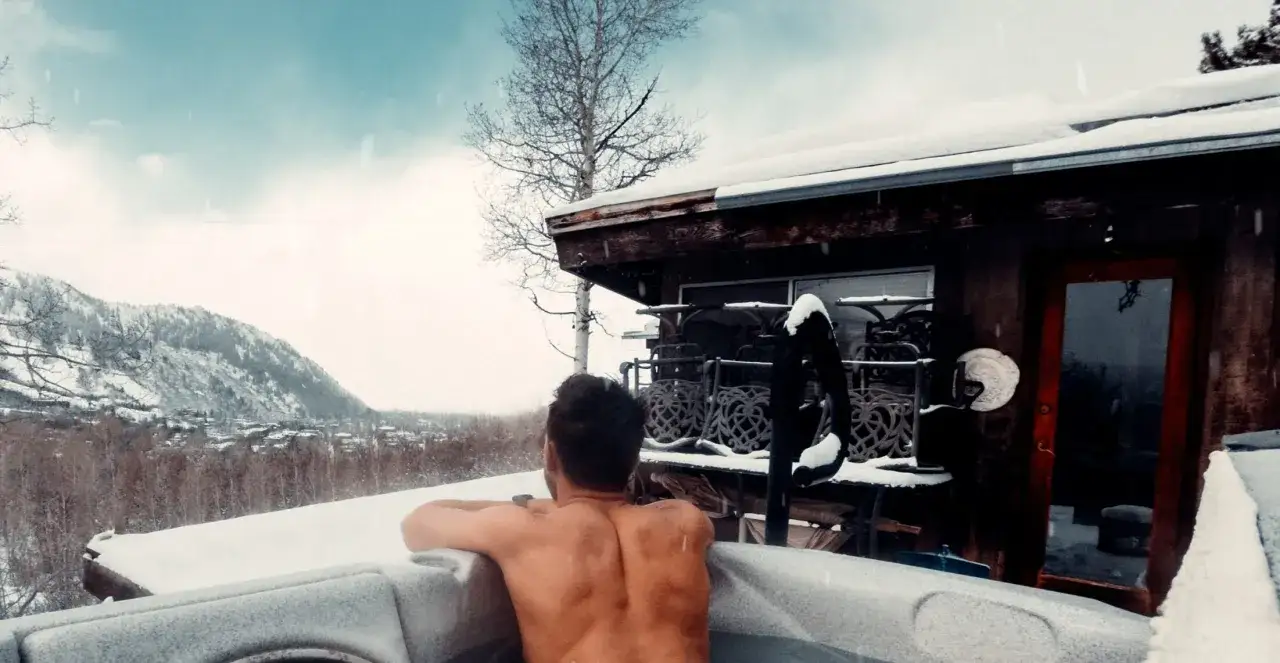 Mężczyzna relaksuje się w jacuzzi ogrodowym lay z spa, podczas gdy zaśnieżone góry i dom tworzą zimową scenerię.