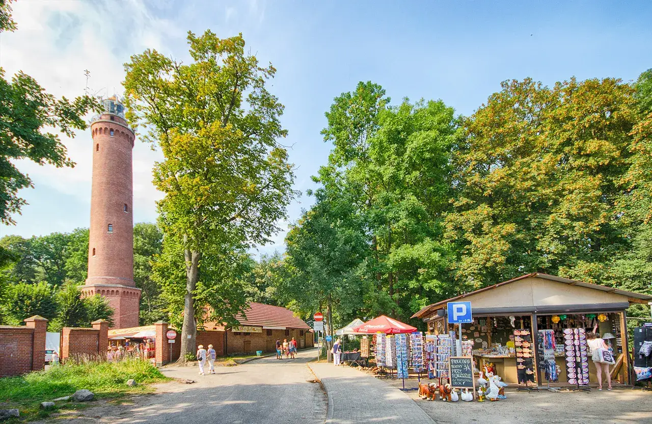 Gąski nad morzem: ceglana latarnia morska, stragany z pamiątkami i zielone drzewa tworzą malowniczy krajobraz.